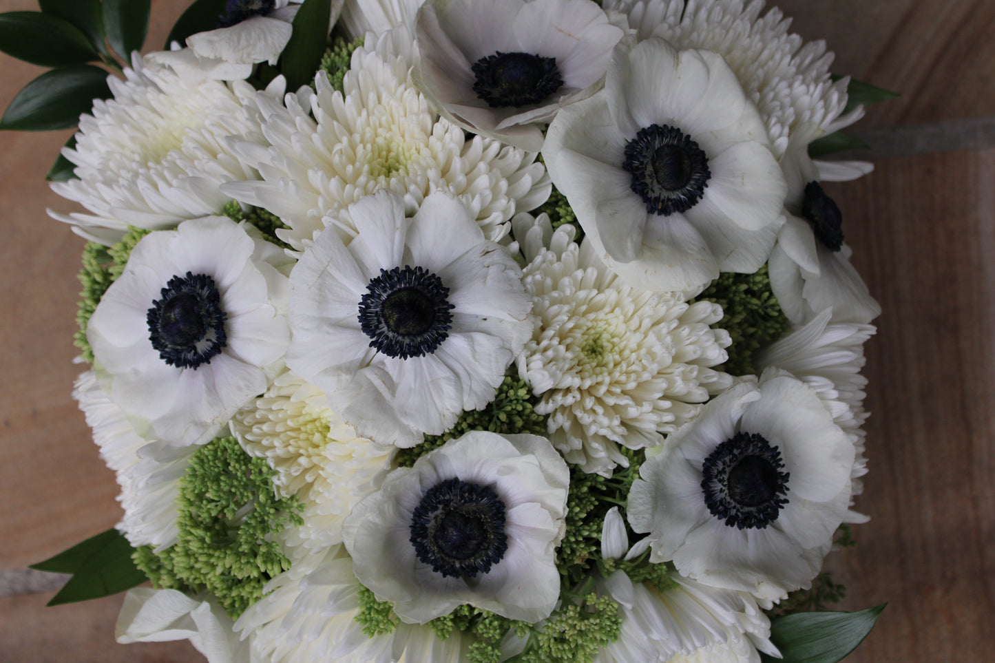 Top view of floral arrangement of black and white anemones, white spider mums, green sedum, and nagi foliage