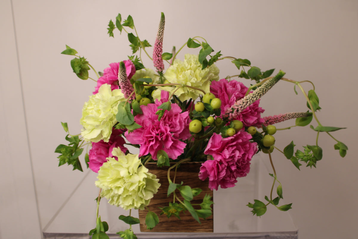 Floral arrangement with pink and green flowers (carnations, veronicas, hypericum berries, and bupleurum) in a wooden cube vase