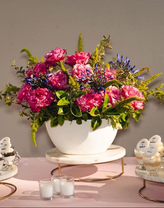 This floral arrangement in a white boat vase features hot pink garden roses, blue agapanthus, green amaranthus, and bright greenery, and sits on a table set with a beautiful light pink tablecloth adorned with candles and petit fours.