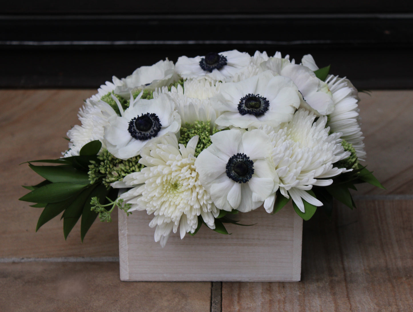 off-white wooden planter box with arrangement of black and white anemones, white spider mums, green sedum, and nagi foliage