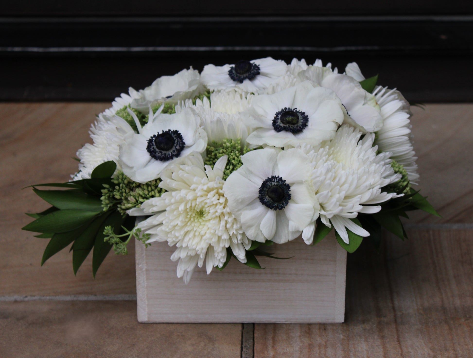 off-white wooden planter box with arrangement of black and white anemones, white spider mums, green sedum, and nagi foliage
