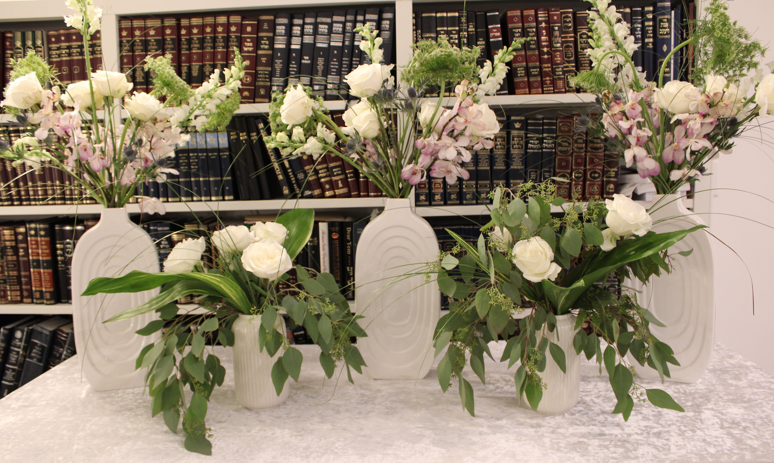 Vort floral arrangements of varying heights. Tall arrangements contain white Tibet roses, mokara orchids, thistles, snapdragons, and Queen Anne's lace. Short arrangements contain white Tibet roses, eucalyptus, and large variegated tropical leaves.