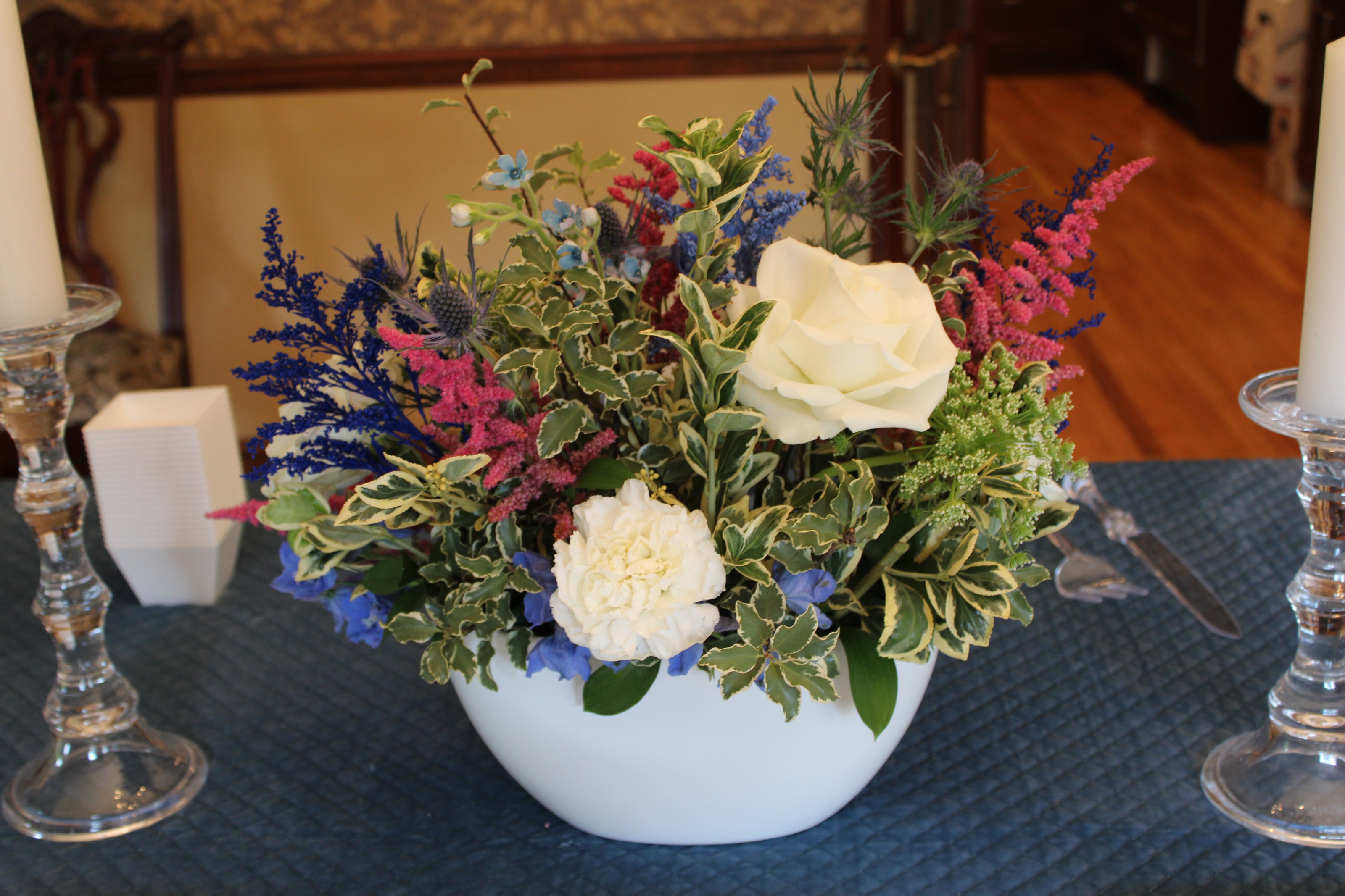 Floral arrangement in a white boat vase on a table with candles and a blue velvet quilted tablecloth. Arrangement includes white carnations, white roses, blue delphinium, hot pink astilbe, dark blue thistle, light blue tweedia, Queen Anne's lace, and variegated greenery.