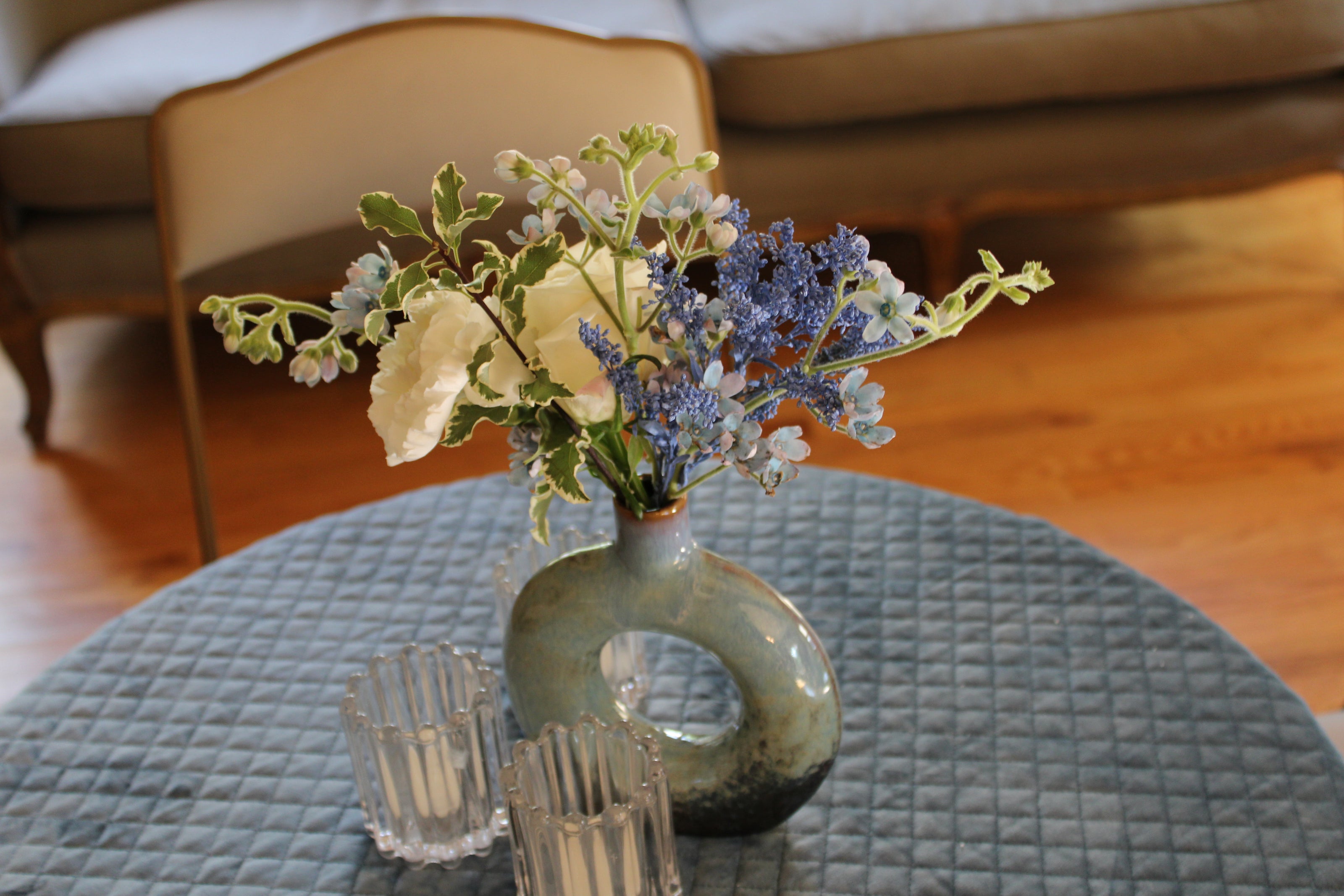 Cool blue/gray vase with tweedia, carnation, rose, painted foliage, and variegated greenery on a light blue velvet quilted tablecloth