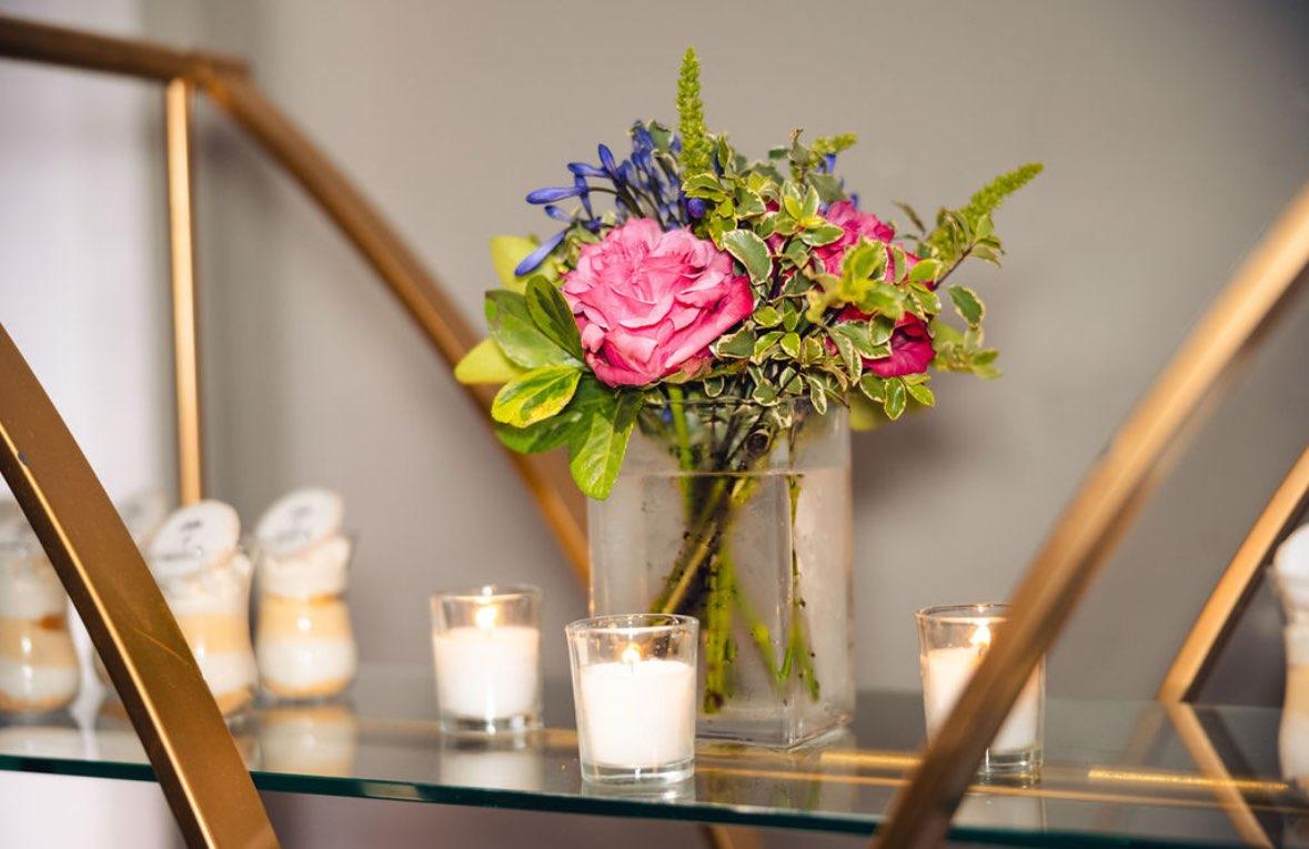 Rectangular glass vase with hot pink garden roses, blue agapanthus, and green amaranthus, surrounded by candles on a glass table