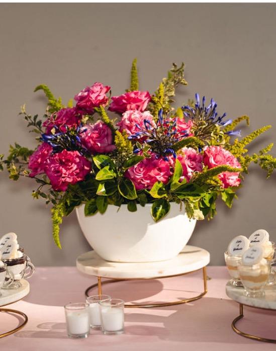 This floral arrangement in a white boat vase features hot pink garden roses, blue agapanthus, green amaranthus, and bright greenery, and sits on a table set with a beautiful light pink tablecloth adorned with candles and petit fours.