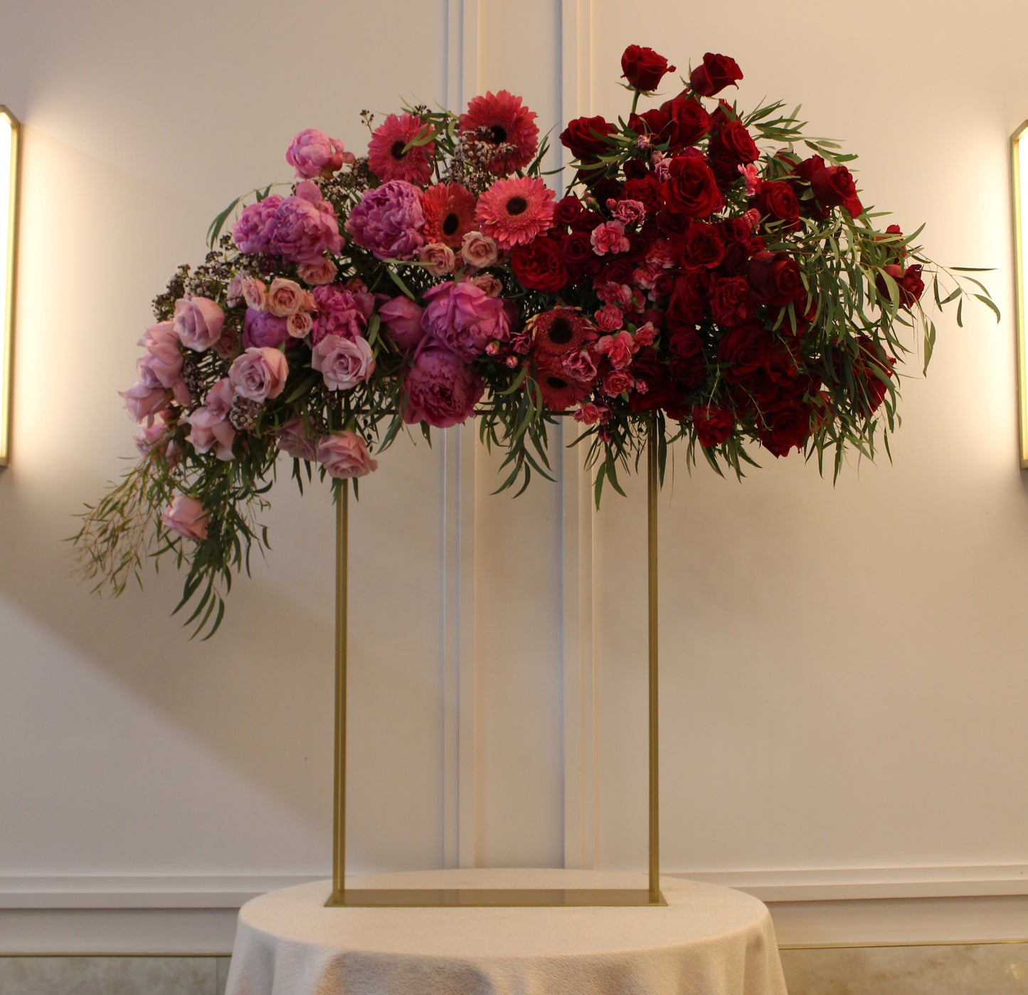 ombre light pink to red large vort floral arrangement on gold stand with peonies, roses, Gerber daisies, carnations, eupatorium, and eucalyptus