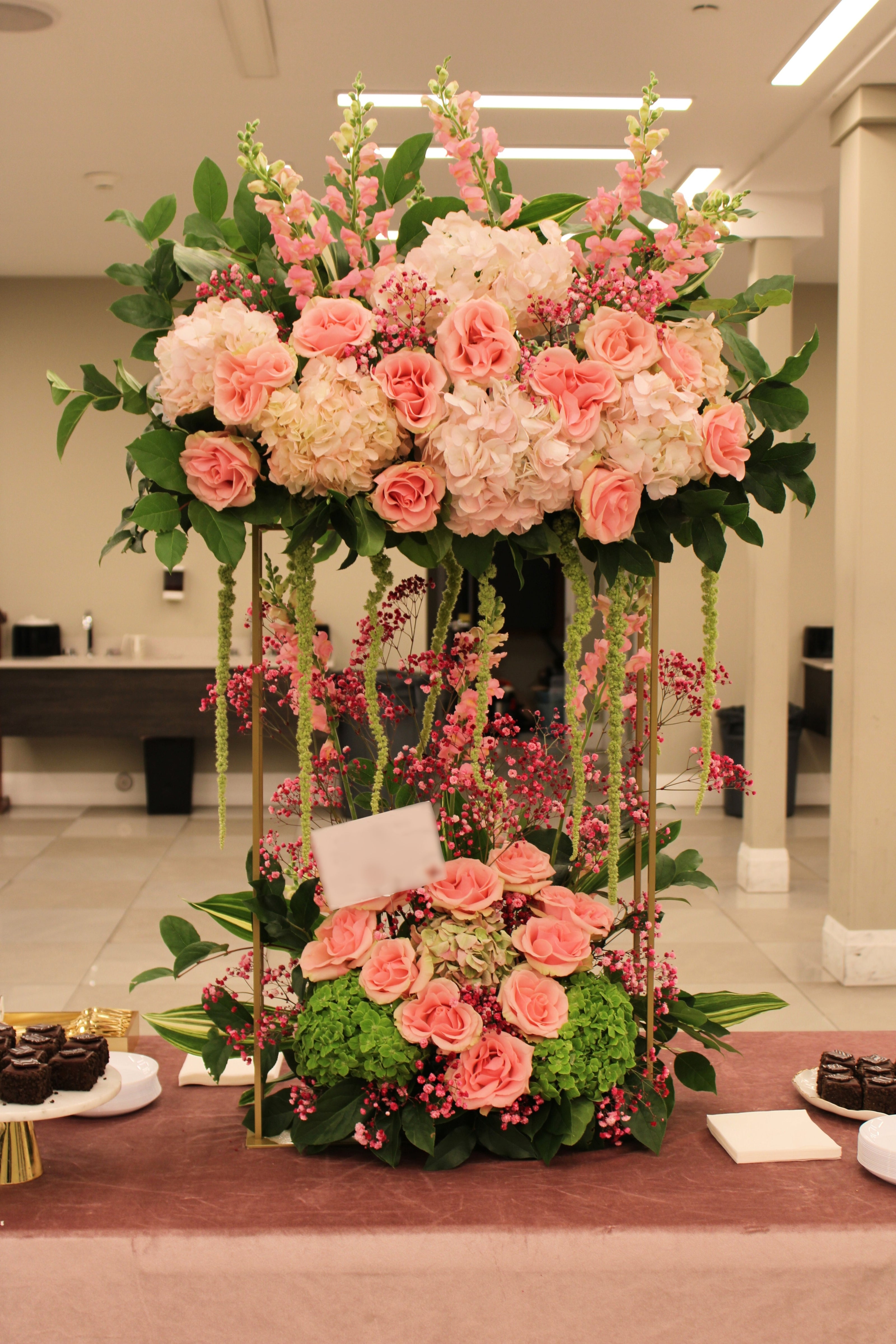 garden style pink and green floral arrangement on gold stand with roses, snapdragons, baby's breath, hydrangeas, hanging amaranthus, lemon leaf, and variegated aspidistra leaves