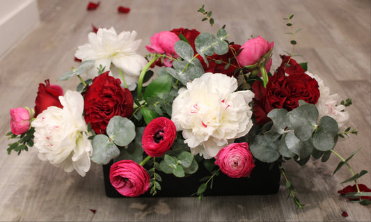 Floral arrangement with white Festiva Maxima peonies, red heart roses, hot pink ranunculus, and mixed greens in a matte black planter vase