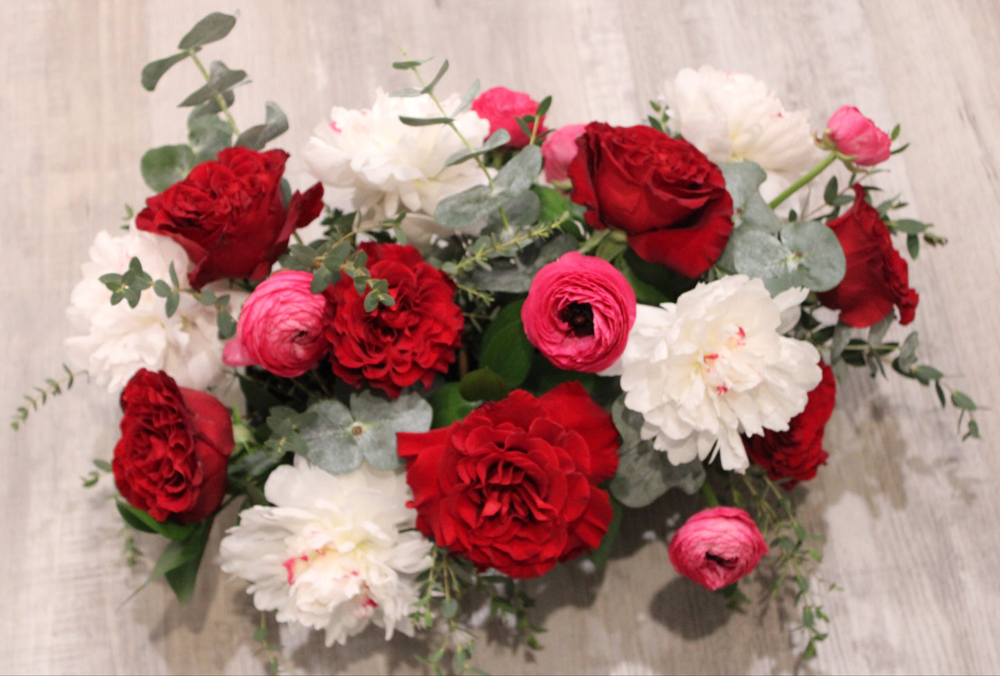 floral arrangement with white Festiva Maxima peonies, red heart roses, hot pink ranunculus, and mixed greens in a black rectangular planter.