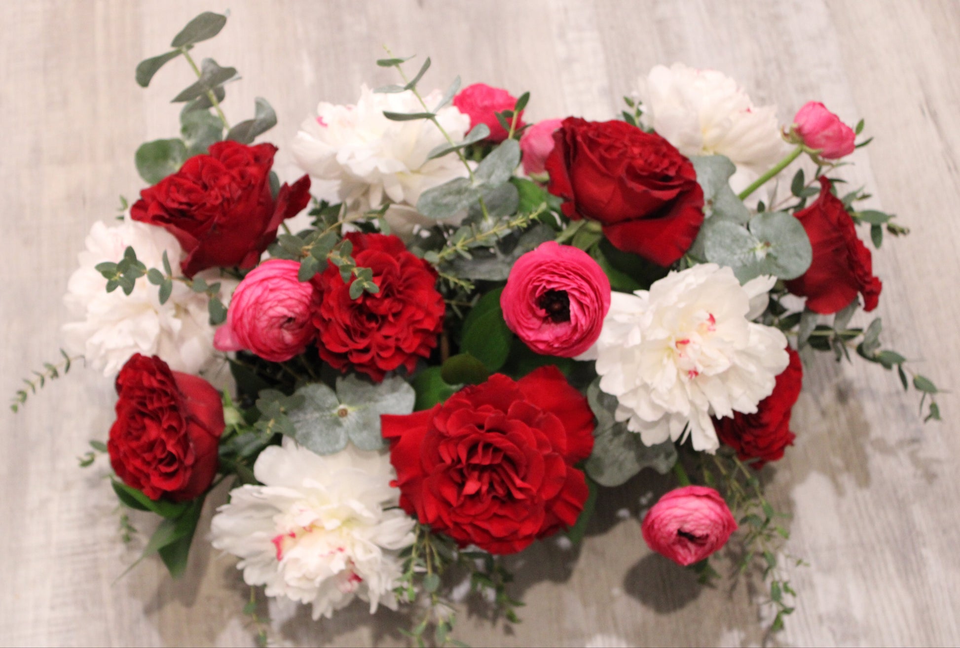 floral arrangement with white Festiva Maxima peonies, red heart roses, hot pink ranunculus, and mixed greens in a black rectangular planter.