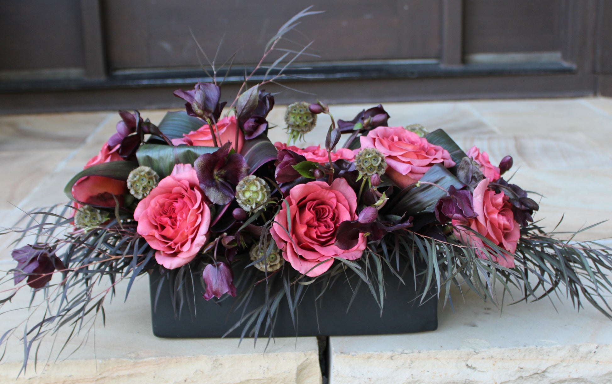 Rectangular black 12" flower vase/planter with Razzmatazz terracotta colored roses, dark purple hellebores, scabiosa pods, red Ti leaves, and agonis foliage